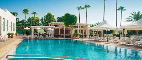 A swimming pool with lounge chairs and umbrellas, surrounded by palm trees and buildings.