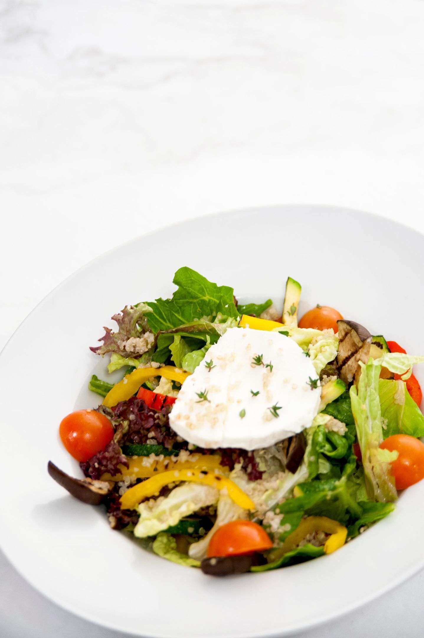 Een wit bord met een salade, diverse groenten, een toefje witte saus en een kers tomaat.