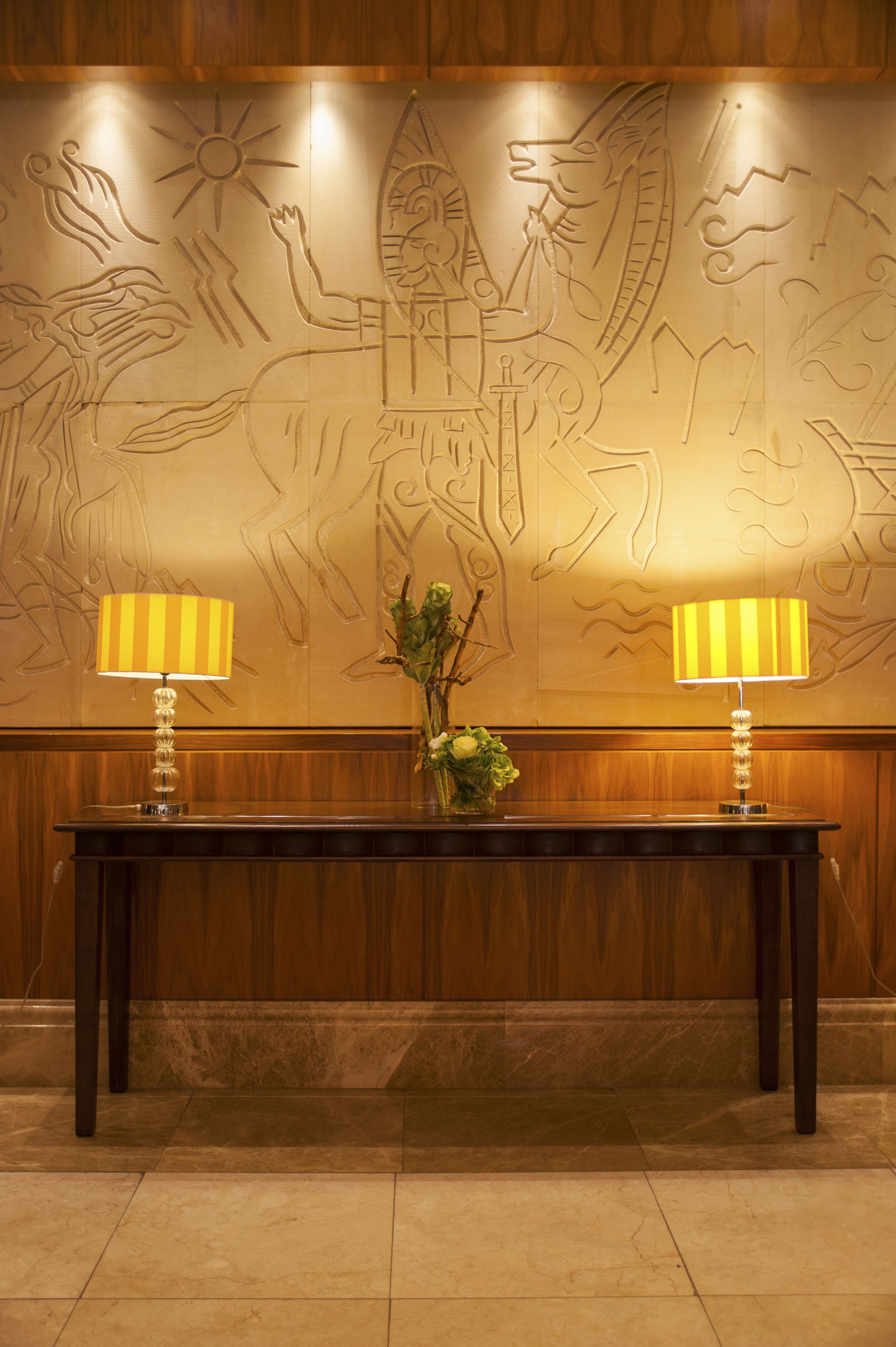 A reception area with a wooden panel wall featuring a relief sculpture, a dark wooden reception desk, two lamps, and a vase with flowers.