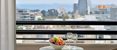 Ein Balkon mit Tisch, auf dem sich eine Obstschale und zwei Weingläser befinden, mit Blick auf eine Stadtlandschaft aus Gebäuden und freiem Meerblick.