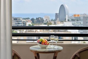 Un balcón con una mesa que sostiene un frutero y dos copas de vino, con vista a un paisaje urbano con edificios y una vista despejada del mar.