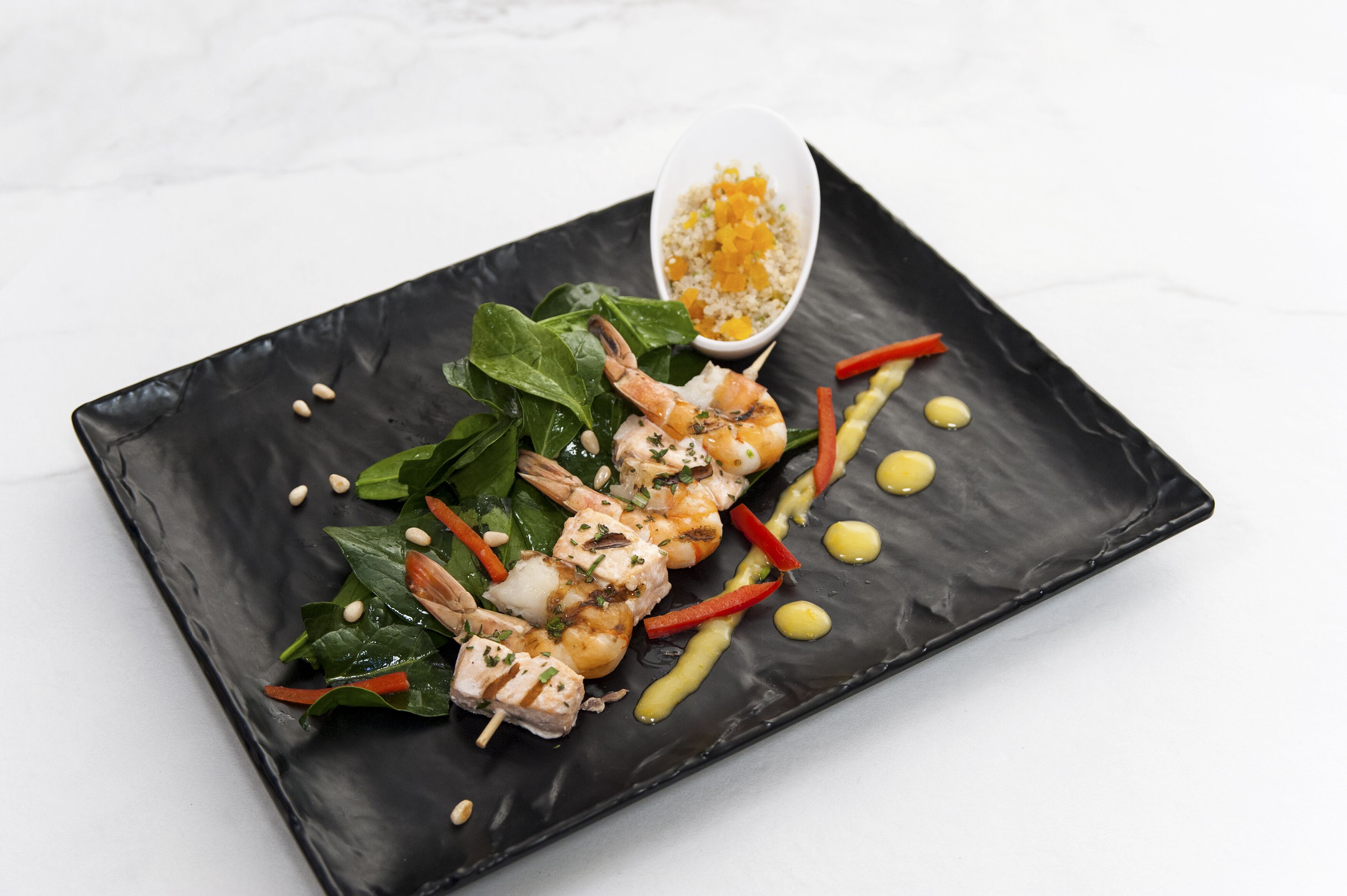 A black rectangular plate with grilled shrimp, spinach, and red bell pepper slices, garnished with sesame seeds and a drizzle of sauce.