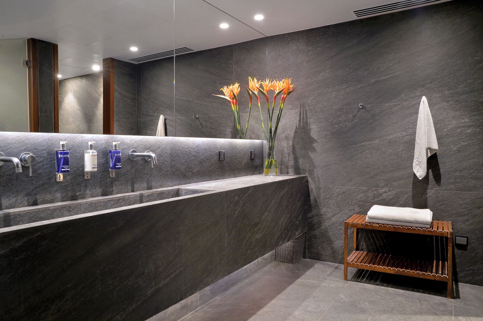 A modern bathroom with a large marble sink, multiple faucets, and a vase with flowers.