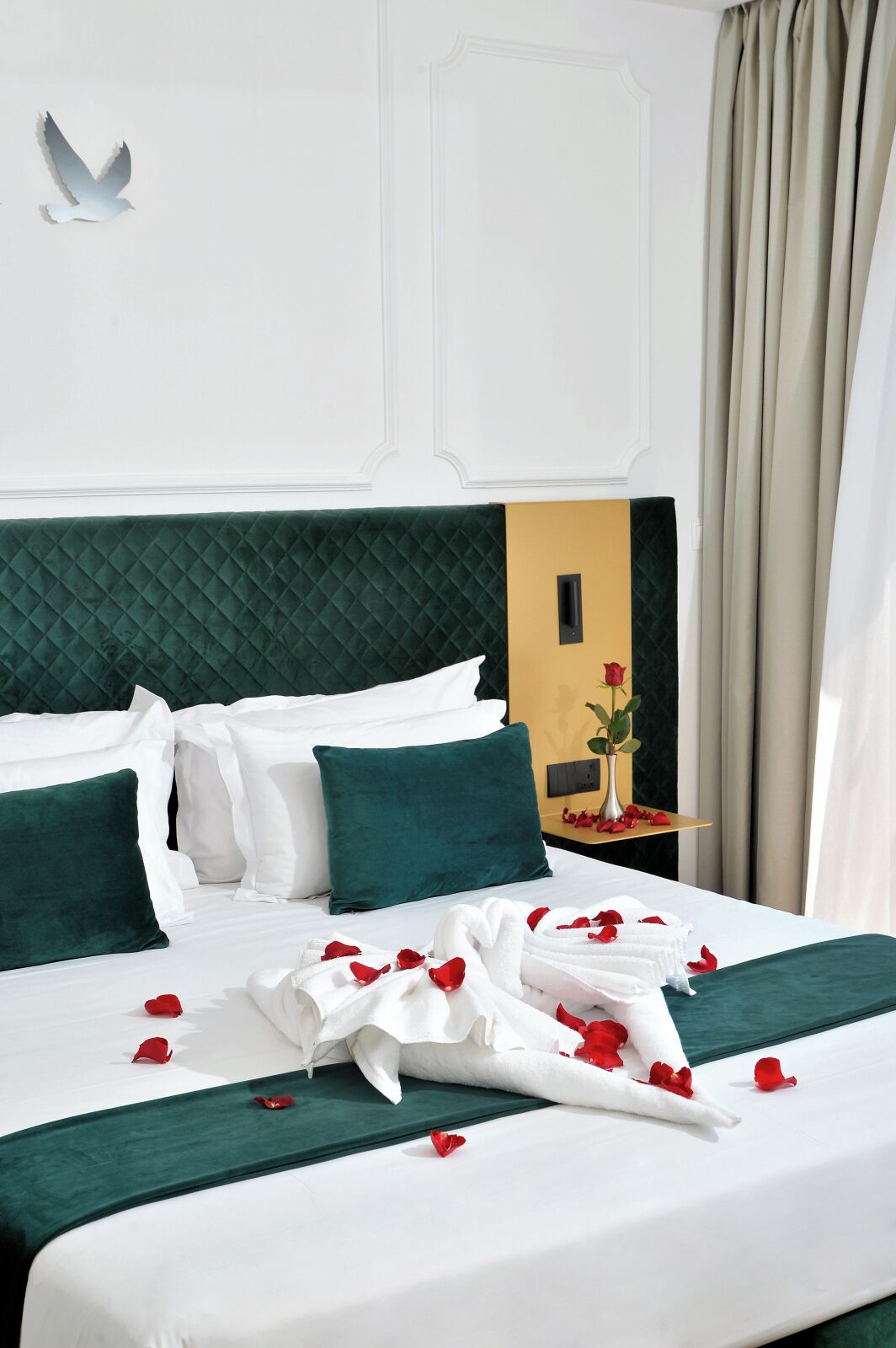 A hotel room with a large bed, green and white bedding, a vase with a single red rose, and a white bird decoration on the wall.