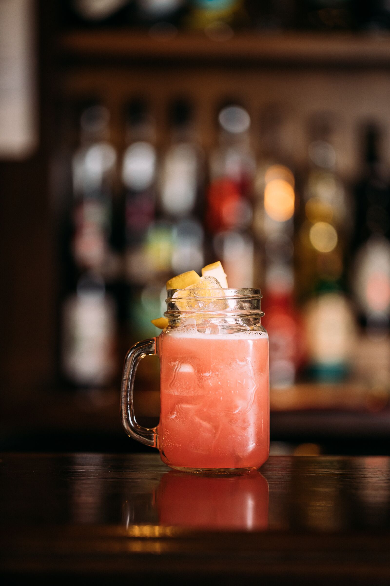 A cocktail in a glass jar with a handle, garnished with a lemon slice, on a reflective surface.