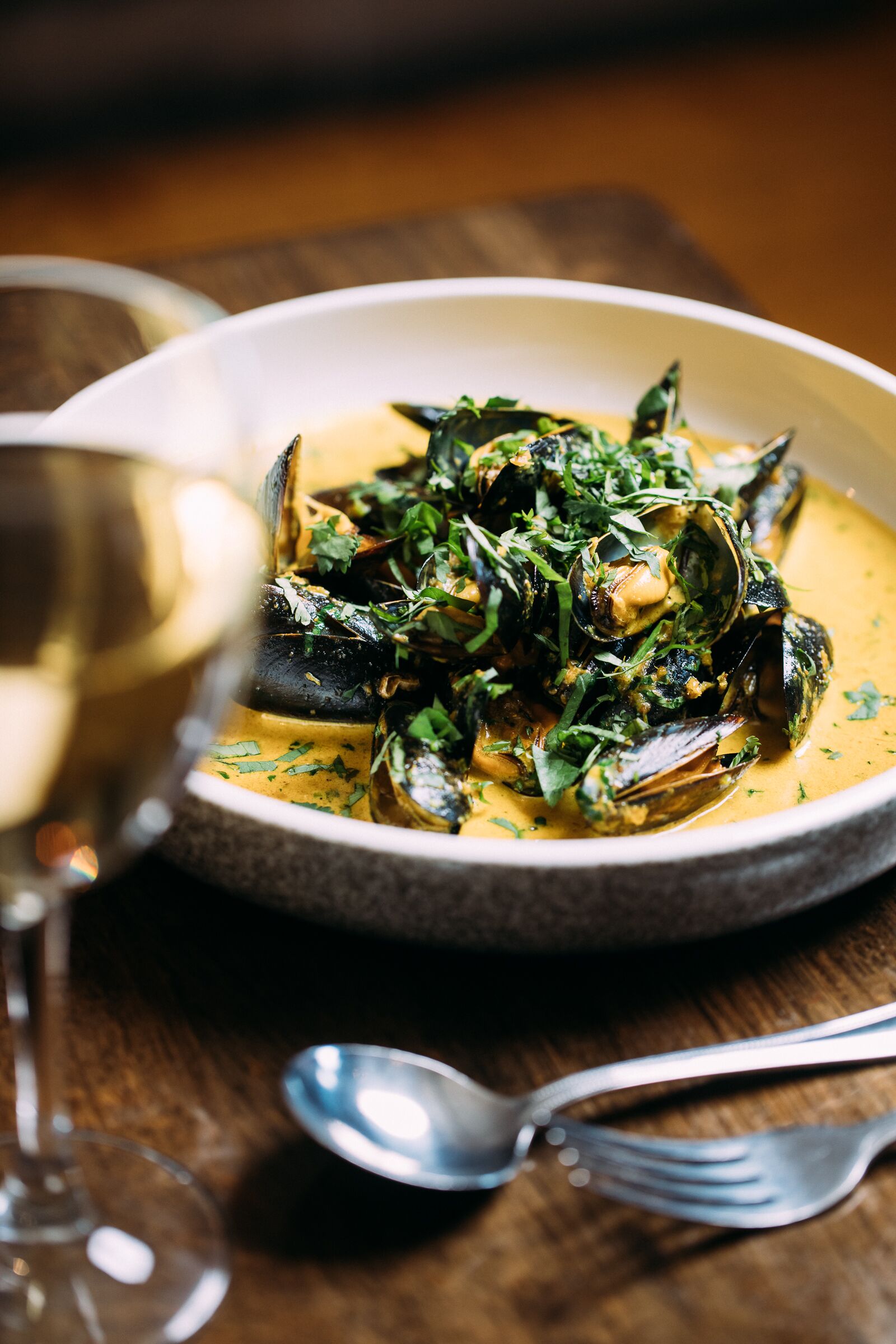 Un bol de mejillones con salsa cremosa, adornado con hierbas, sobre una mesa de madera con una copa de vino y cubiertos.