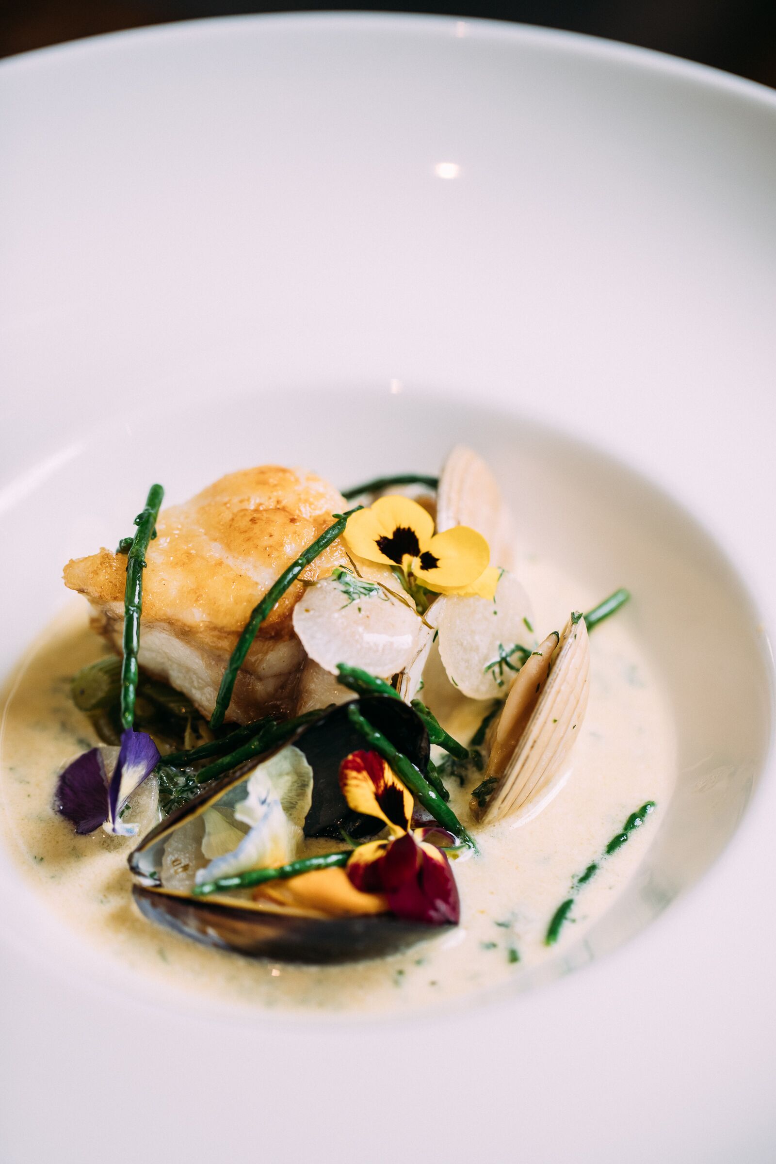 Un plato blanco con un guiso de mariscos, incluyendo mejillones y un trozo de pescado, adornado con flores y hierbas.