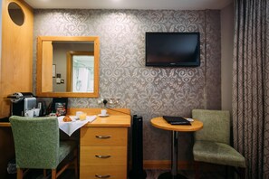 Ett hotellrum med ett skrivbord i trä, en grön klädd stol, ett litet runt bord, en platt-TV som är monterad på väggen och tapeter med ett mönster.
