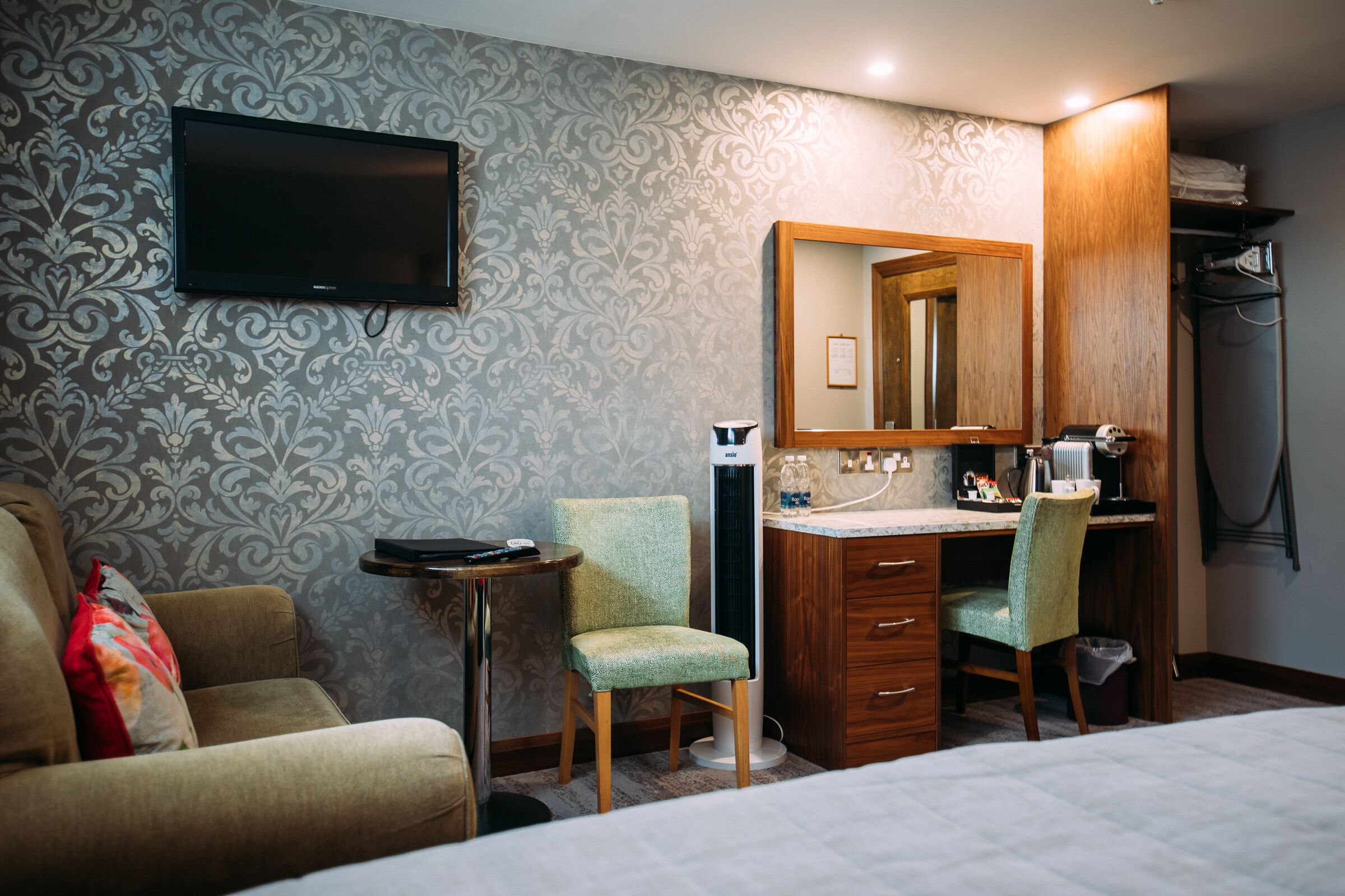 Quarto de hotel com cama, escrivaninha com cadeira, televisão na parede e um espelho grande acima da escrivaninha.