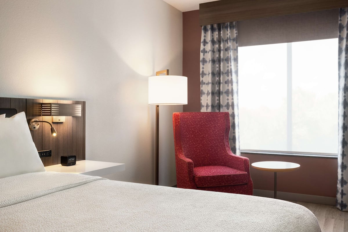 Een hotelkamer met een bed, een rode fauteuil, een klein tafeltje, een lamp en een raam met gordijnen.