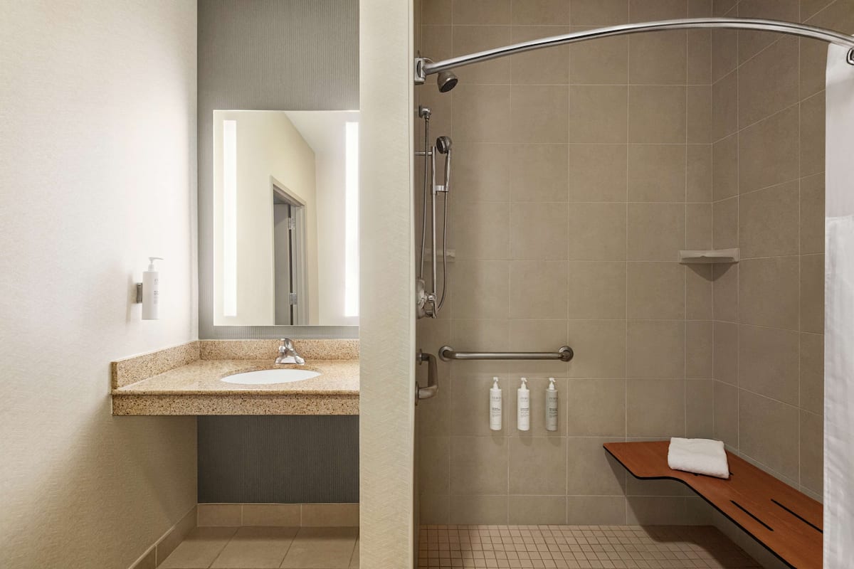 Een badkamer met een wastafel met granieten blad, een spiegel, een douche met een gebogen glazen deur en een houten bankje.