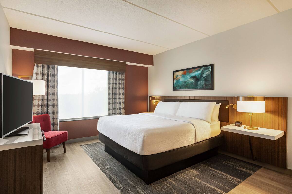 Een hotelkamer met een bed, een bureau met een televisie, een rode stoel, een nachtkastje met een lamp en een raam met gordijnen.