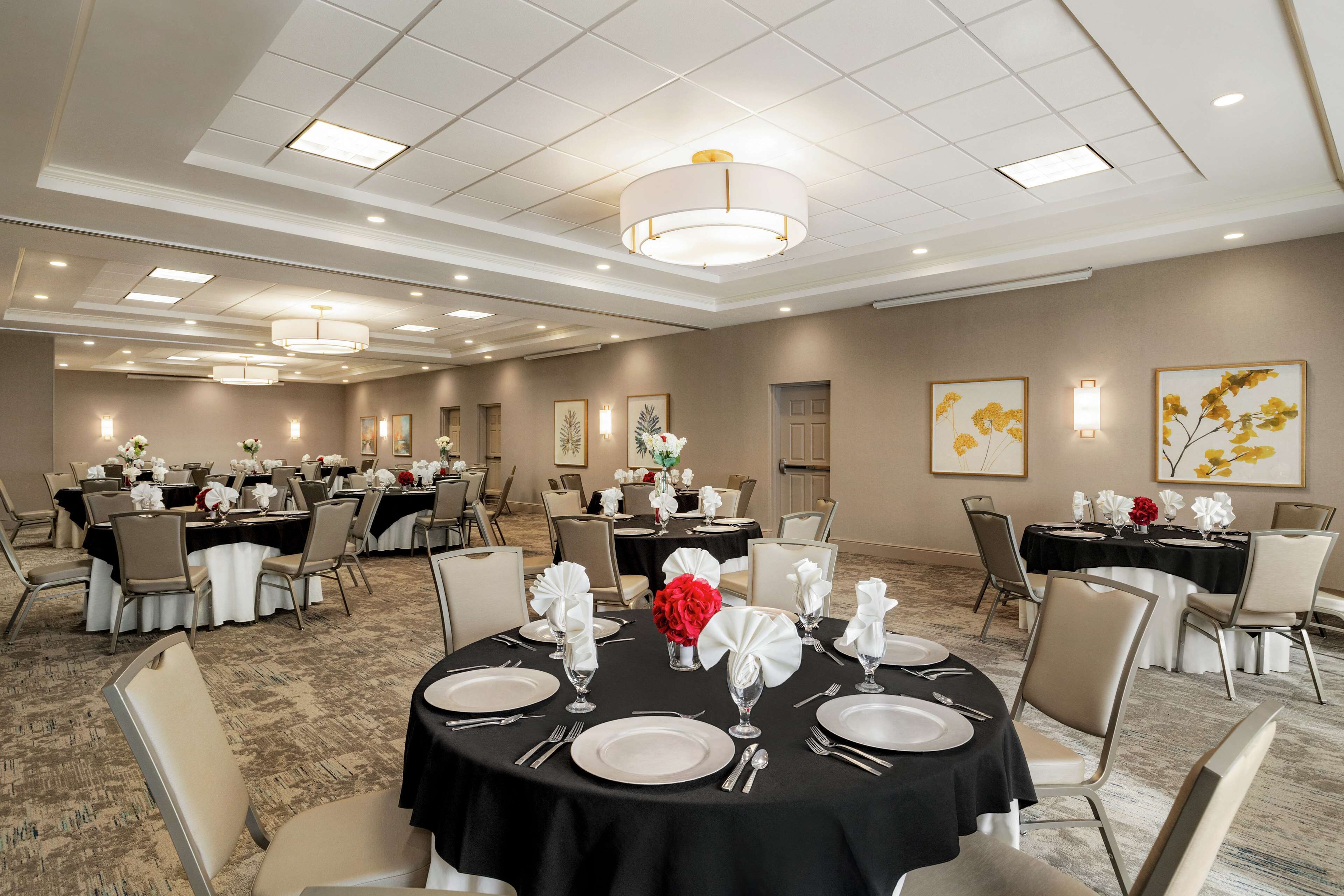Un salón de banquetes con mesas redondas dispuestas para el servicio de comidas, con manteles blancos, cubiertos y flores rojas.