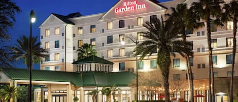 Un hotel de varios pisos con un llamativo letrero de "Hilton Garden Inn", una entrada con techo verde y palmeras en primer plano.