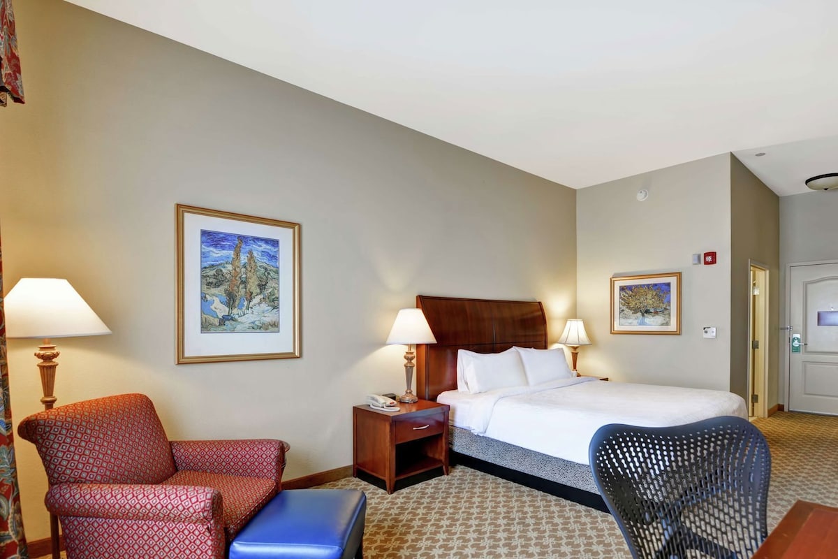 A hotel room with a bed, two bedside tables, a chair, a small table, and two wall-mounted lamps.