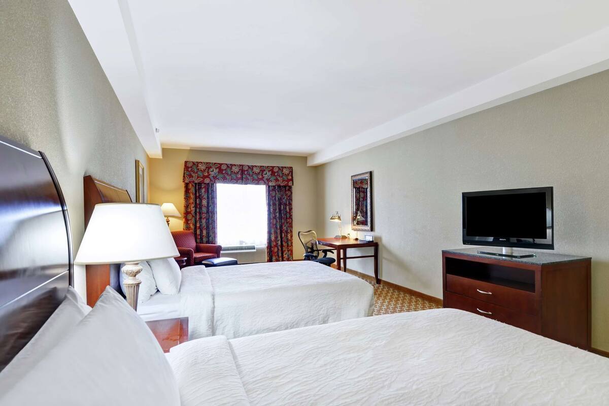 A hotel room with two beds, a desk with a chair, a television, and a window with curtains.