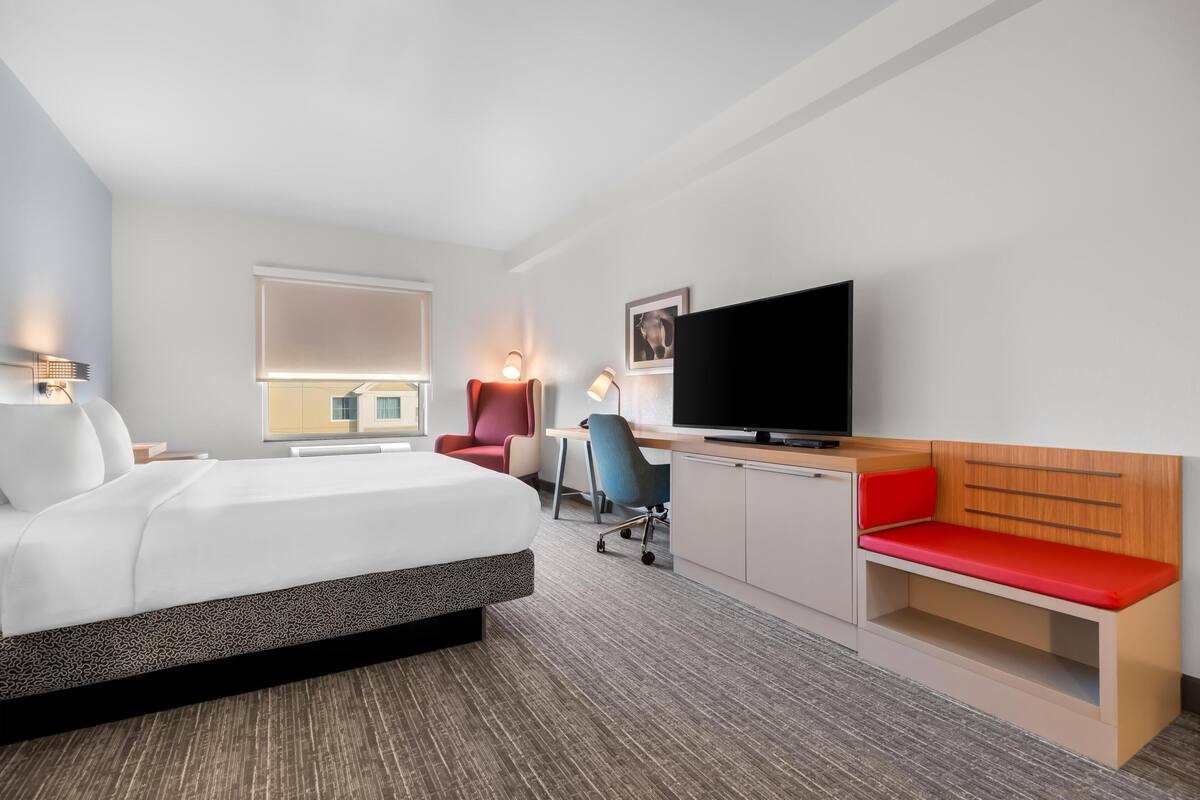 A modern hotel room with a bed, desk, chair, TV, and seating area.