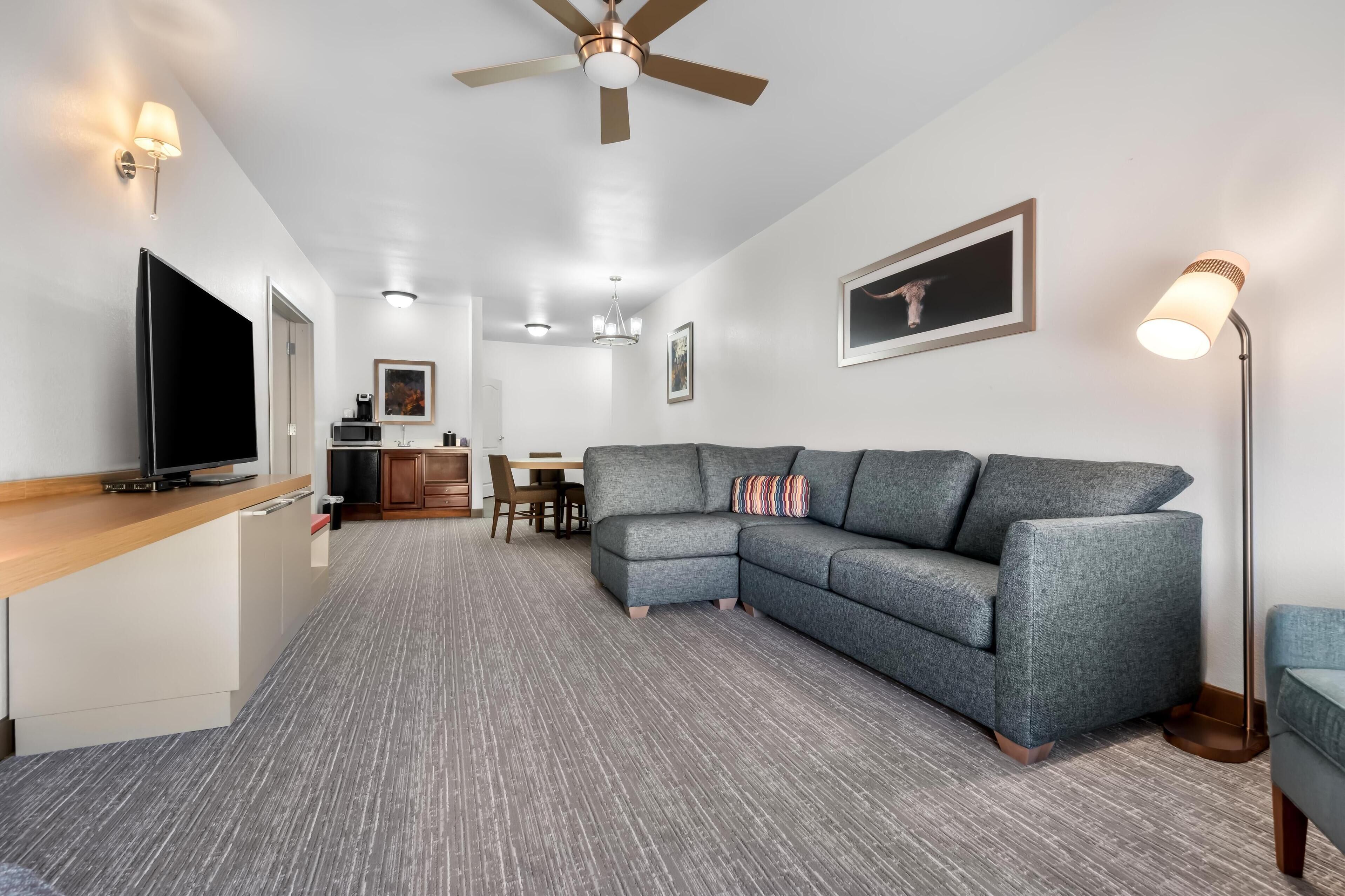 A modern living room with a grey sectional sofa, a flat-screen TV, a ceiling fan, and a dining area in the background.
