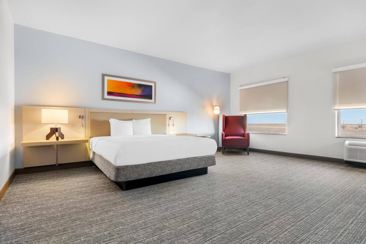 A hotel room with a bed, a desk with a lamp, a chair, and a painting on the wall.
