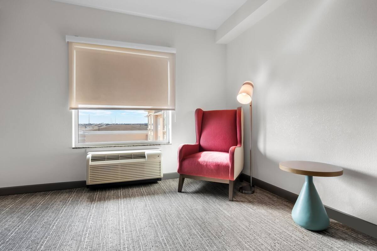 A hotel room with a large window, a red armchair, a floor lamp, and a small round table.