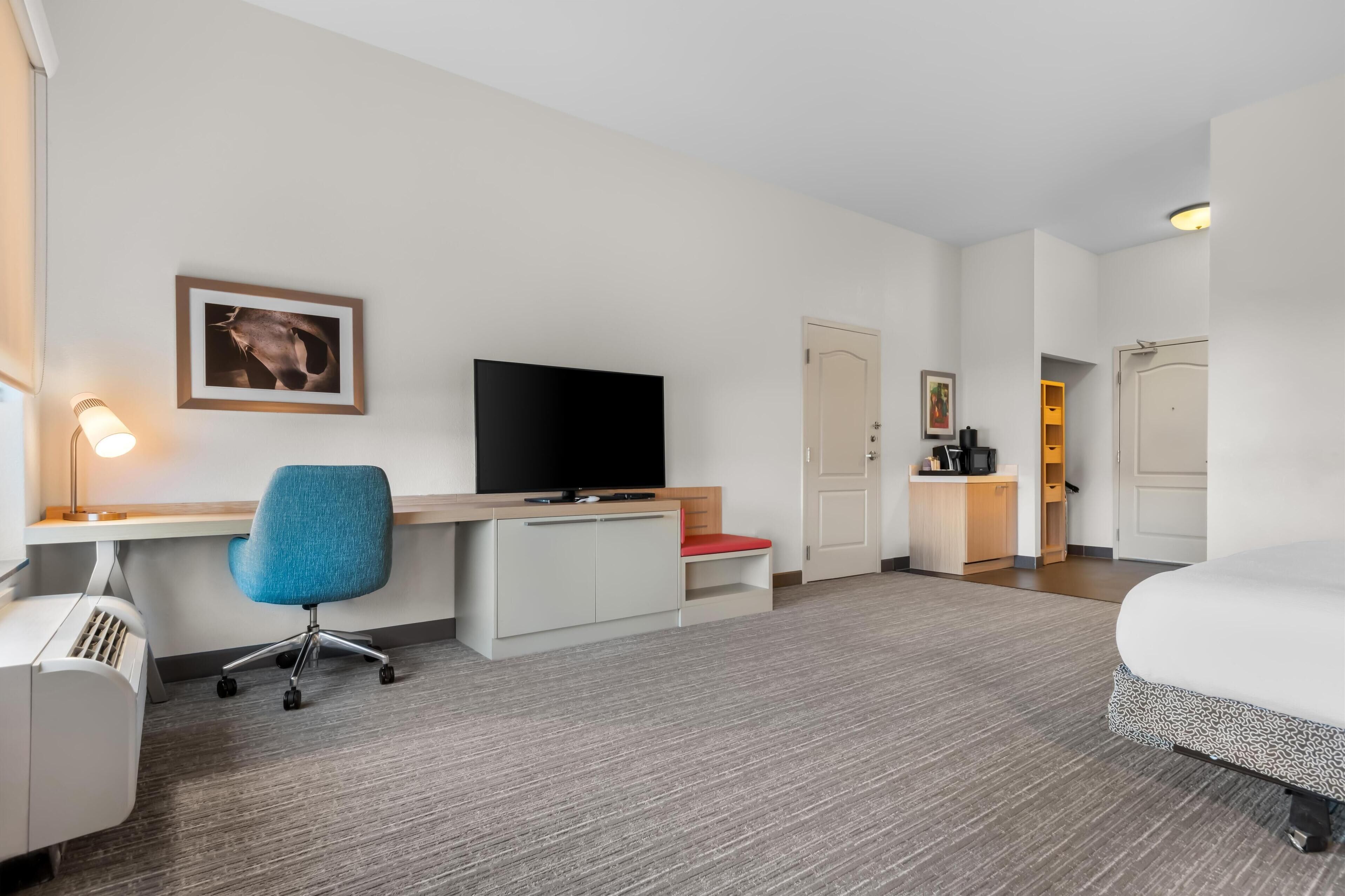 A hotel room with a bed, desk, chair, TV, and kitchenette.