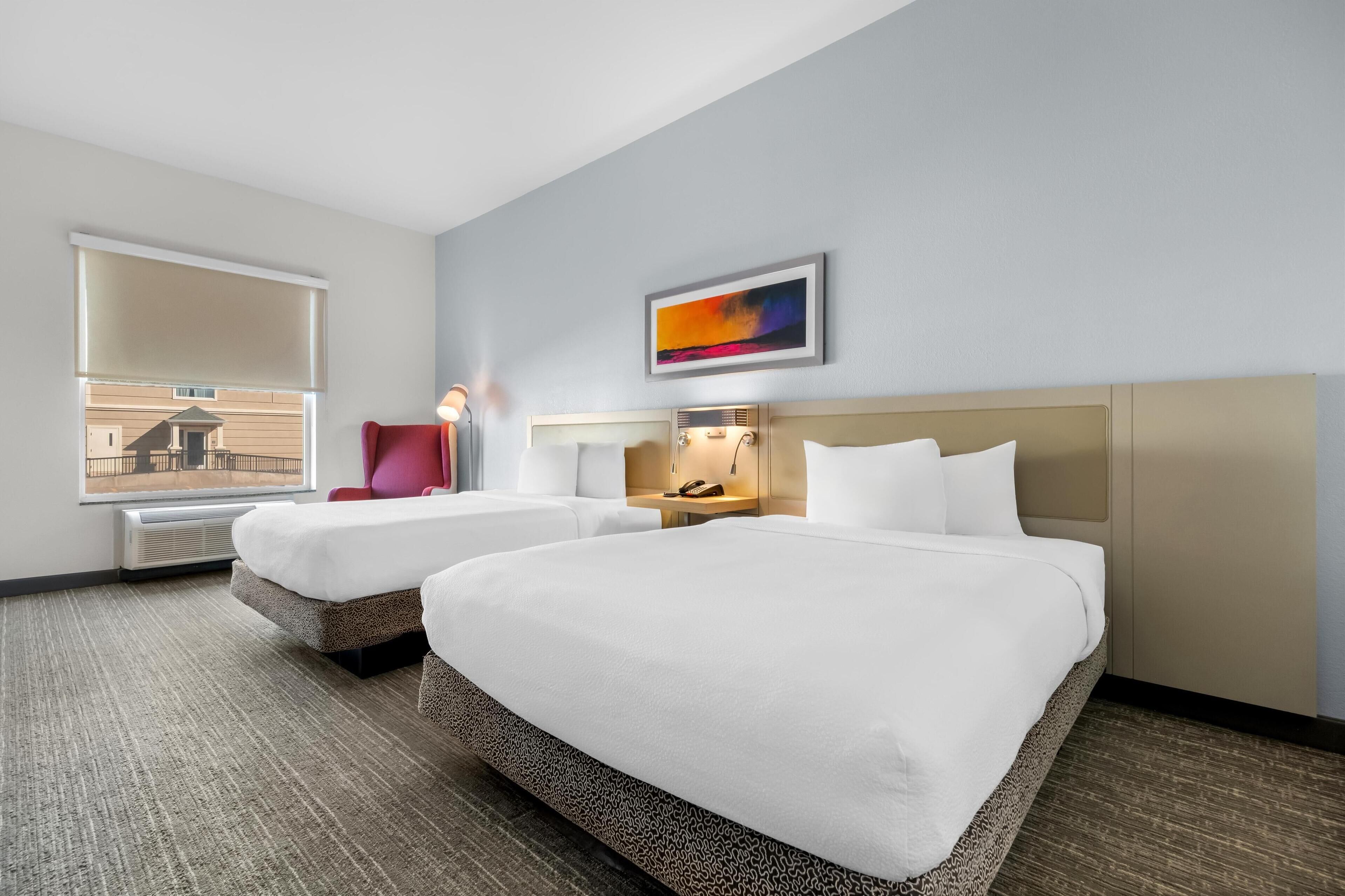 Una habitación de hotel con dos camas, una silla roja, una mesita de noche con lámpara, un cuadro en la pared y una ventana con persianas.