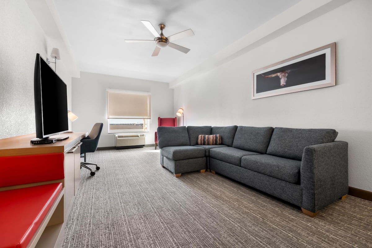 A modern hotel room with a grey sectional sofa, a red bench, a flat-screen TV, a desk, a ceiling fan, and a framed picture on the wall.