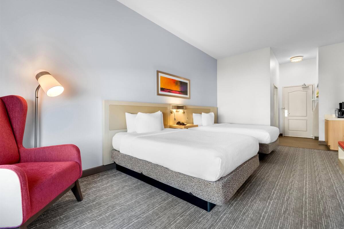 A hotel room with a bed, a red chair, a floor lamp, and a painting on the wall.