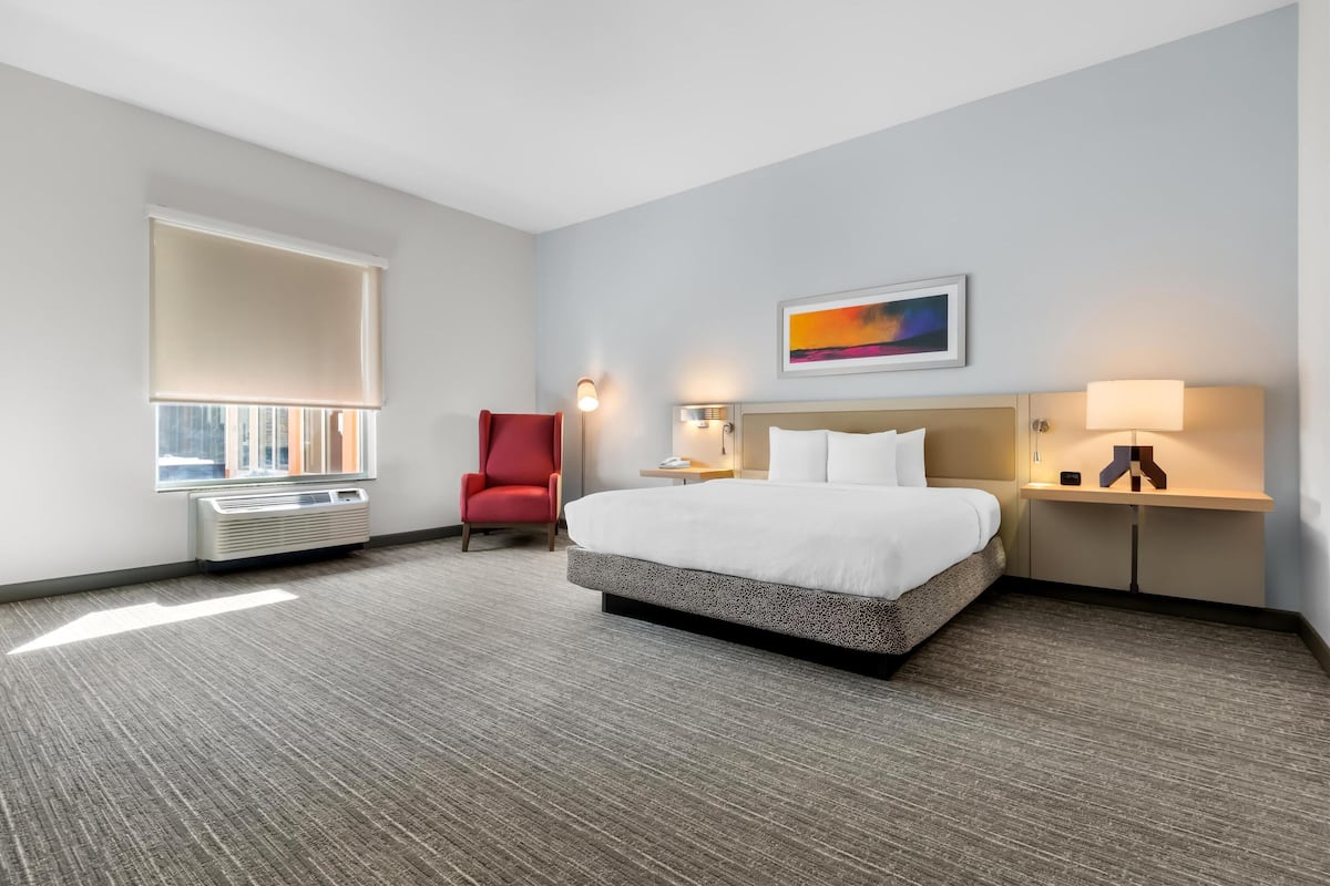 A hotel room with a large bed, a red chair, a window with blinds, a painting on the wall, and a nightstand with a lamp.