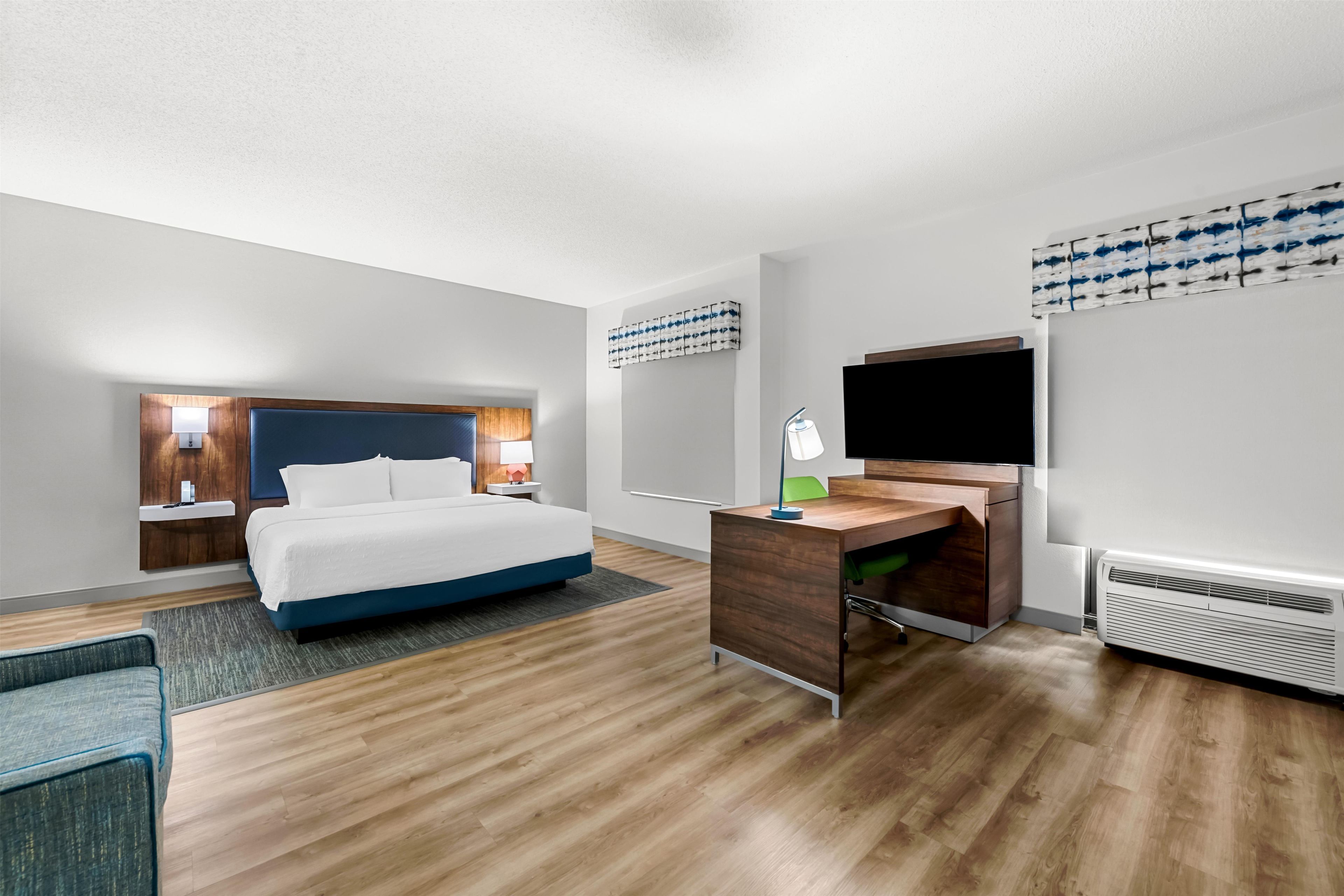A hotel room with a bed, desk, chair, bedside tables, wall art, and a flat-screen TV.
