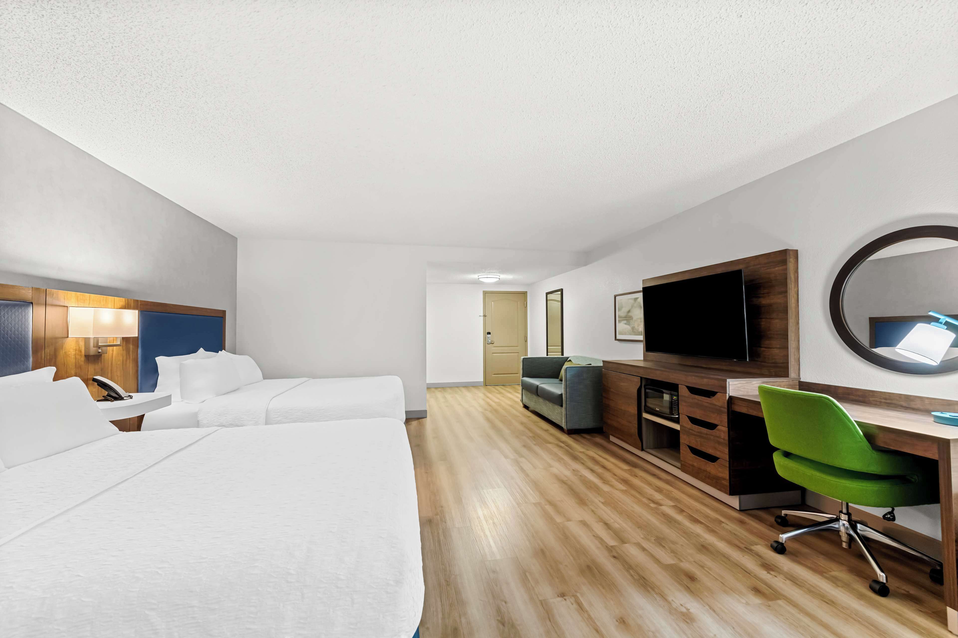 A hotel room with two beds, a desk with a chair, a TV, a mirror, and a door leading to another room.