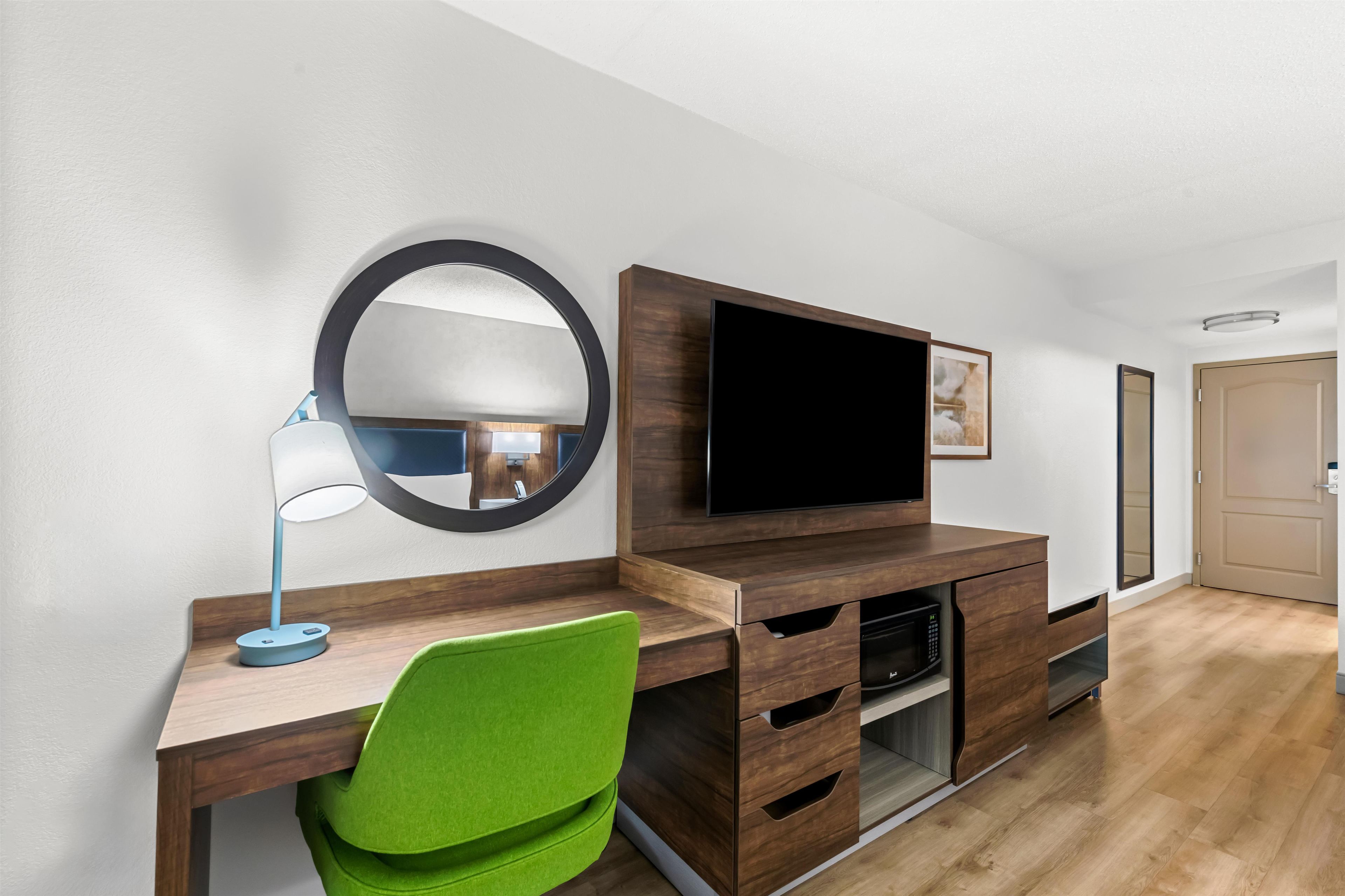 Una habitación moderna con un escritorio de madera, una silla verde, un espejo redondo, un televisor de pantalla plana y un mueble empotrado para entretenimiento.
