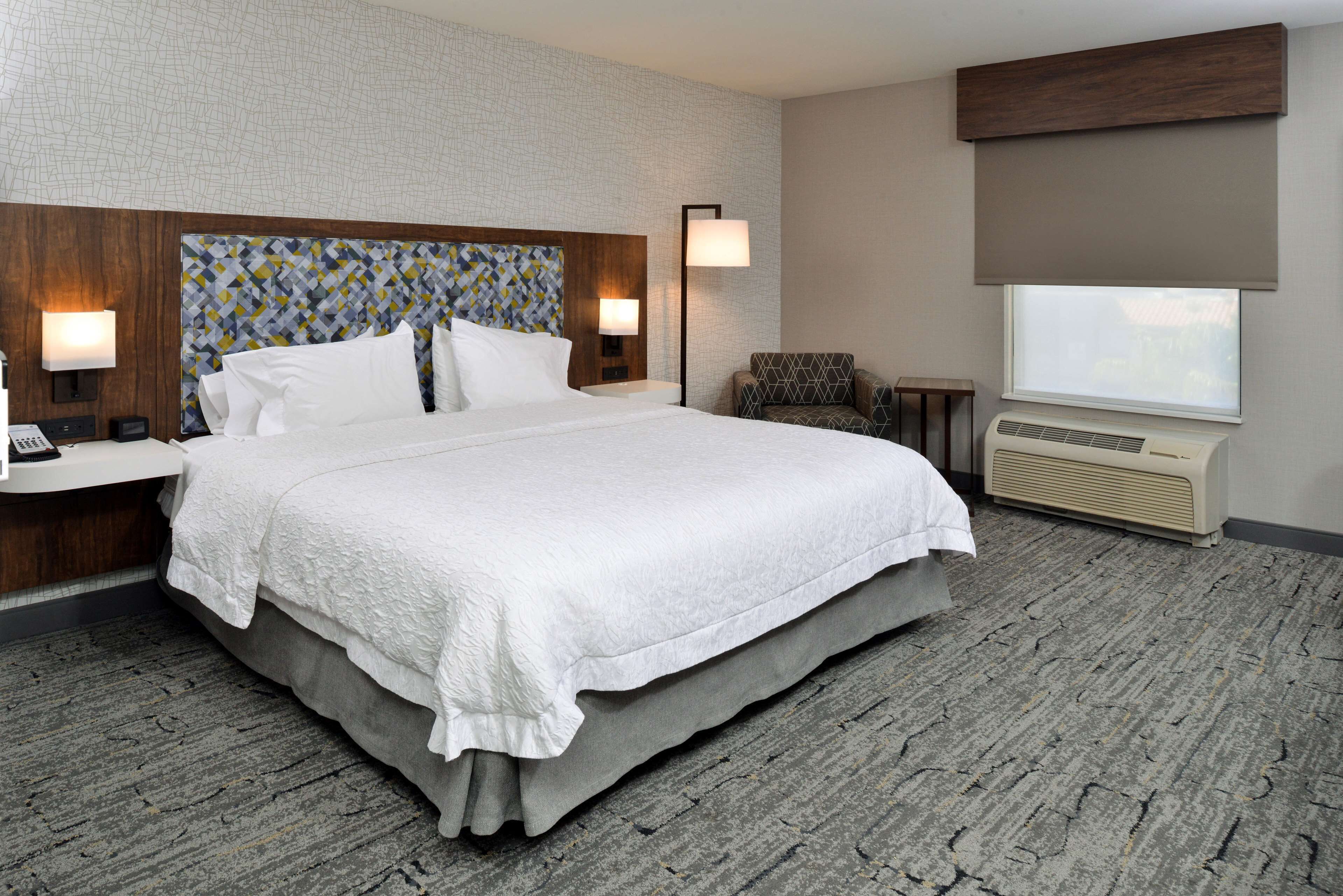 Hotelkamer met een groot bed, twee nachtkastjes, een stoel, een bureau, een lamp, een raam met zonwering en een wandairconditioner.