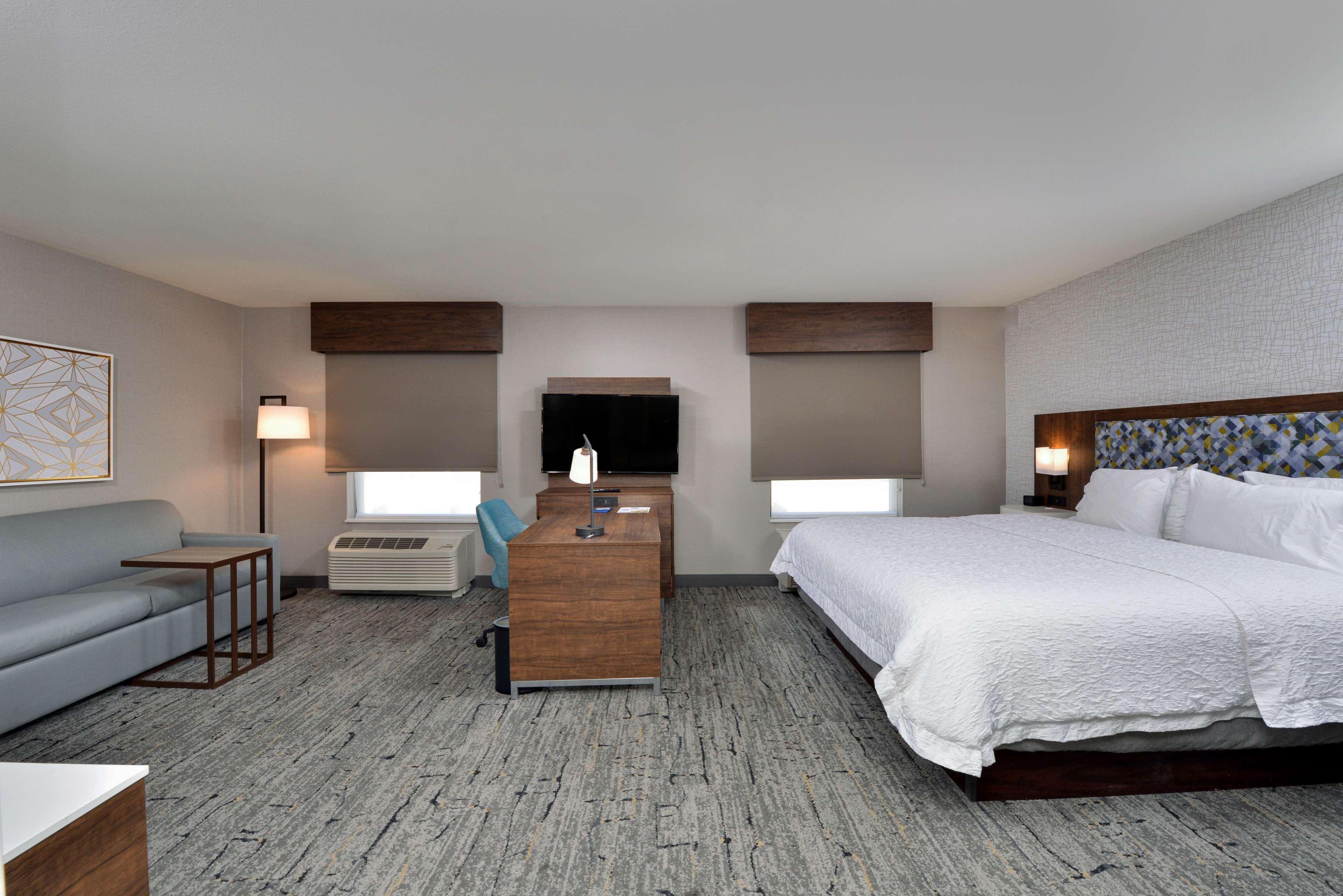 A hotel room with a bed, a desk, a chair, a sofa, a TV, and a lamp.