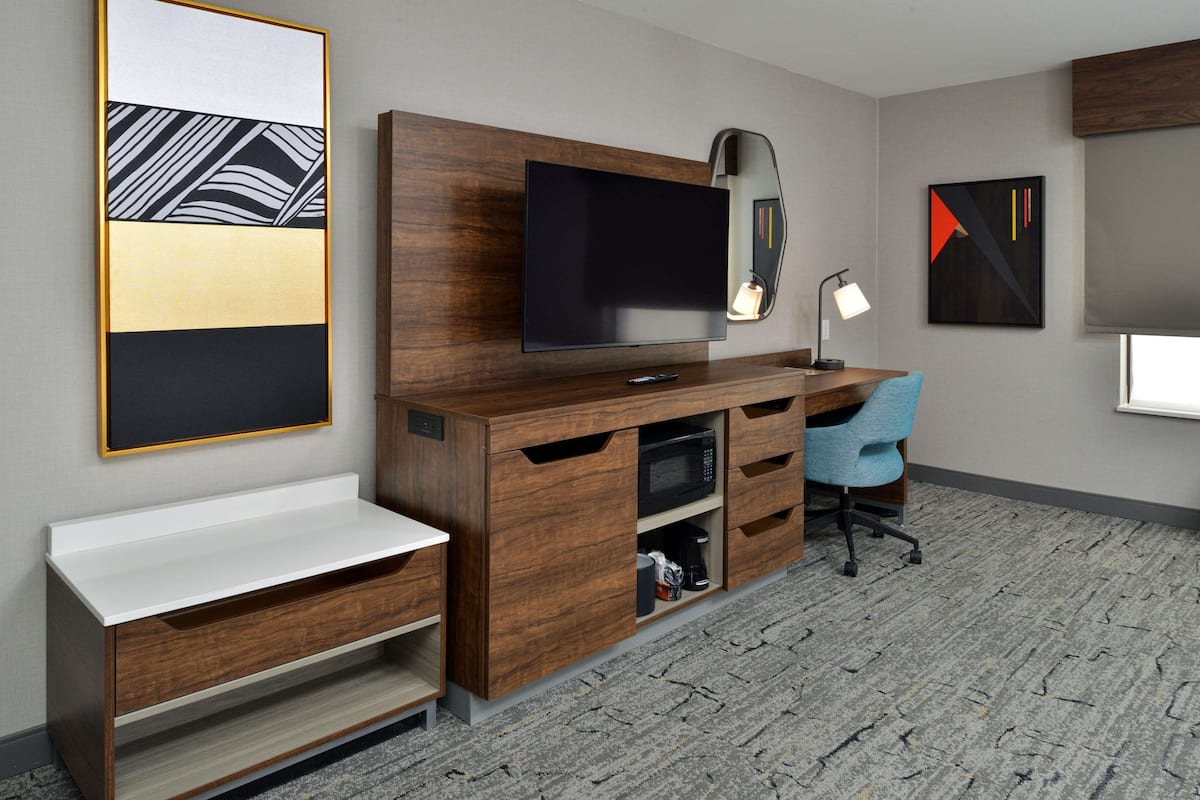 A modern hotel room with a wooden desk, a flat-screen TV, a blue office chair, and artwork on the walls.