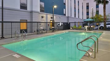 A swimming pool with a metal ladder, surrounded by a black fence, and a building with large windows in the background.