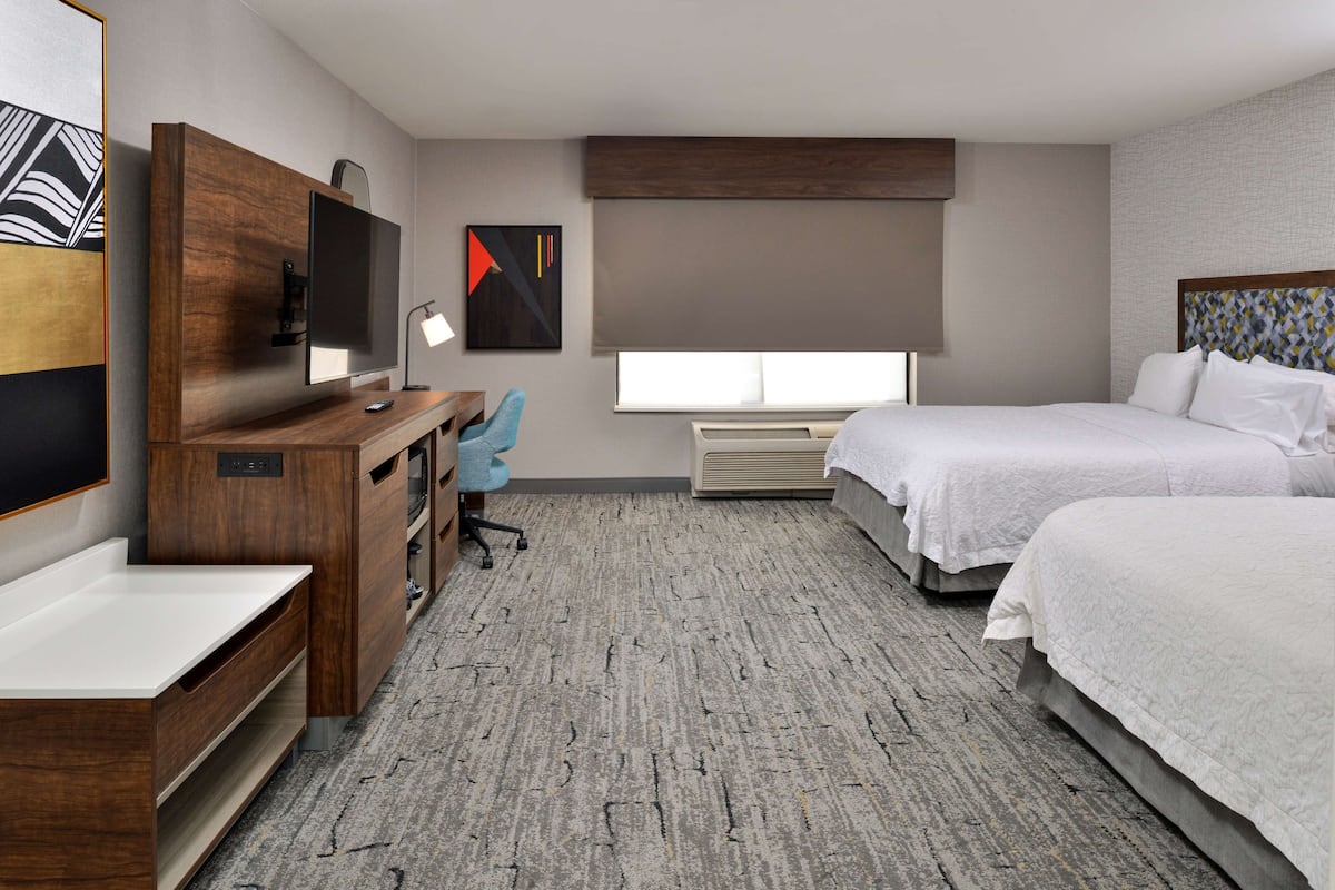 A hotel room with a wooden desk, a television, a desk lamp, a chair, a bed, and a window with a view.