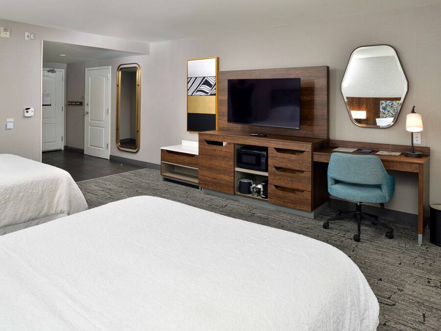 A hotel room with two beds, a TV, a desk with a chair, a mirror, and a lamp.