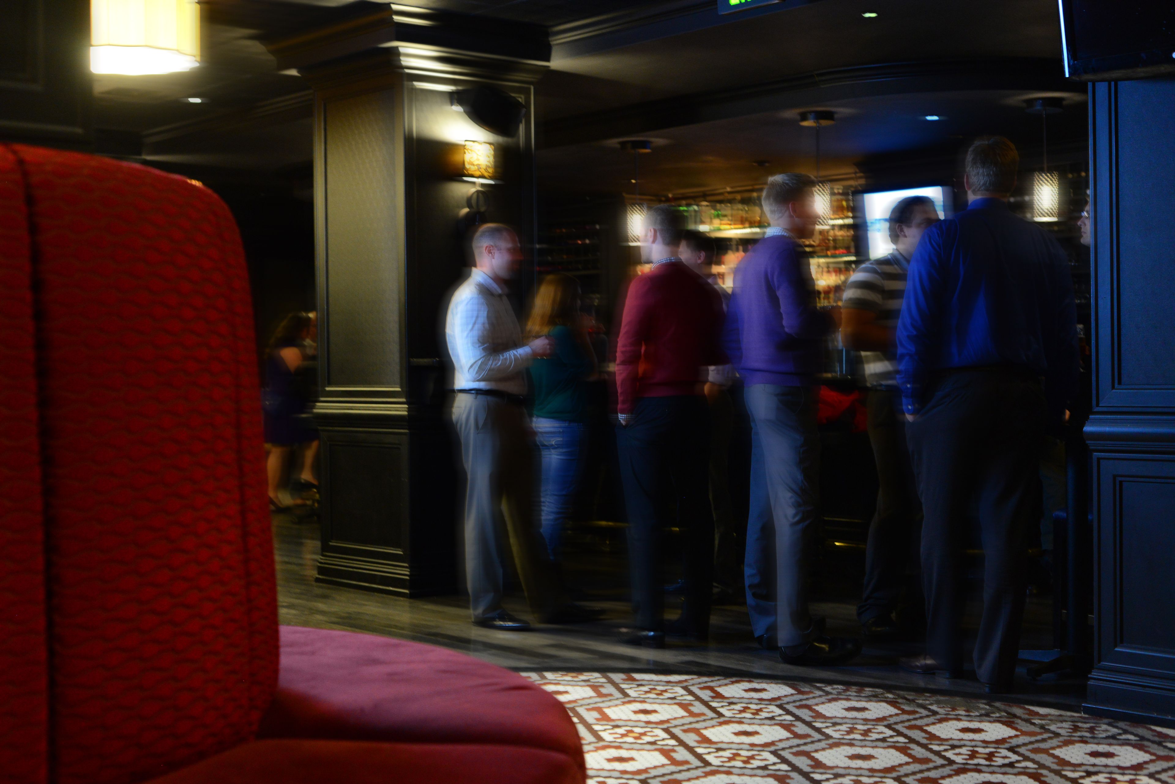A bar area with a patterned floor, red seating, and people standing and conversing.