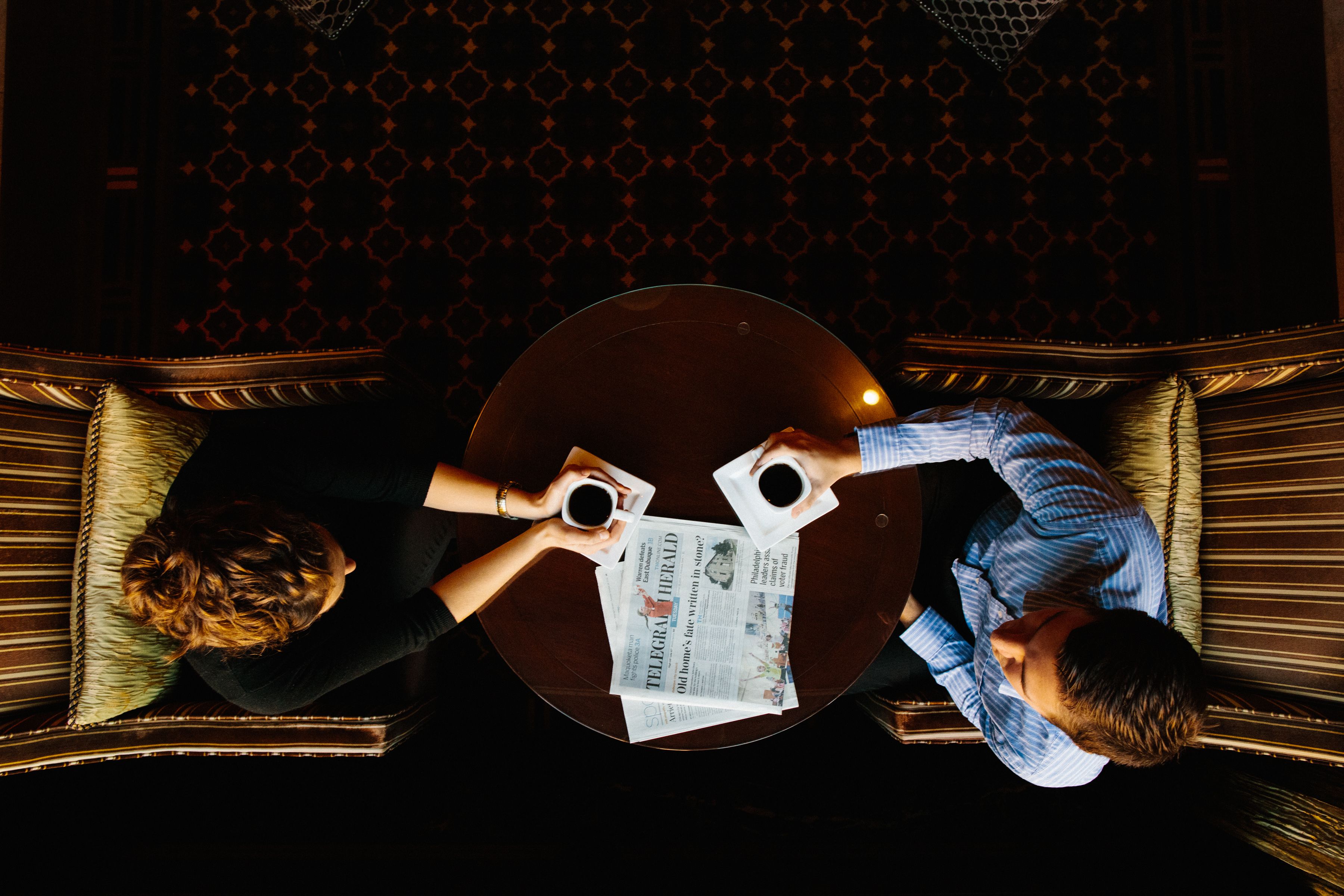 A round table with two people seated, each holding a cup. A newspaper is placed on the table.