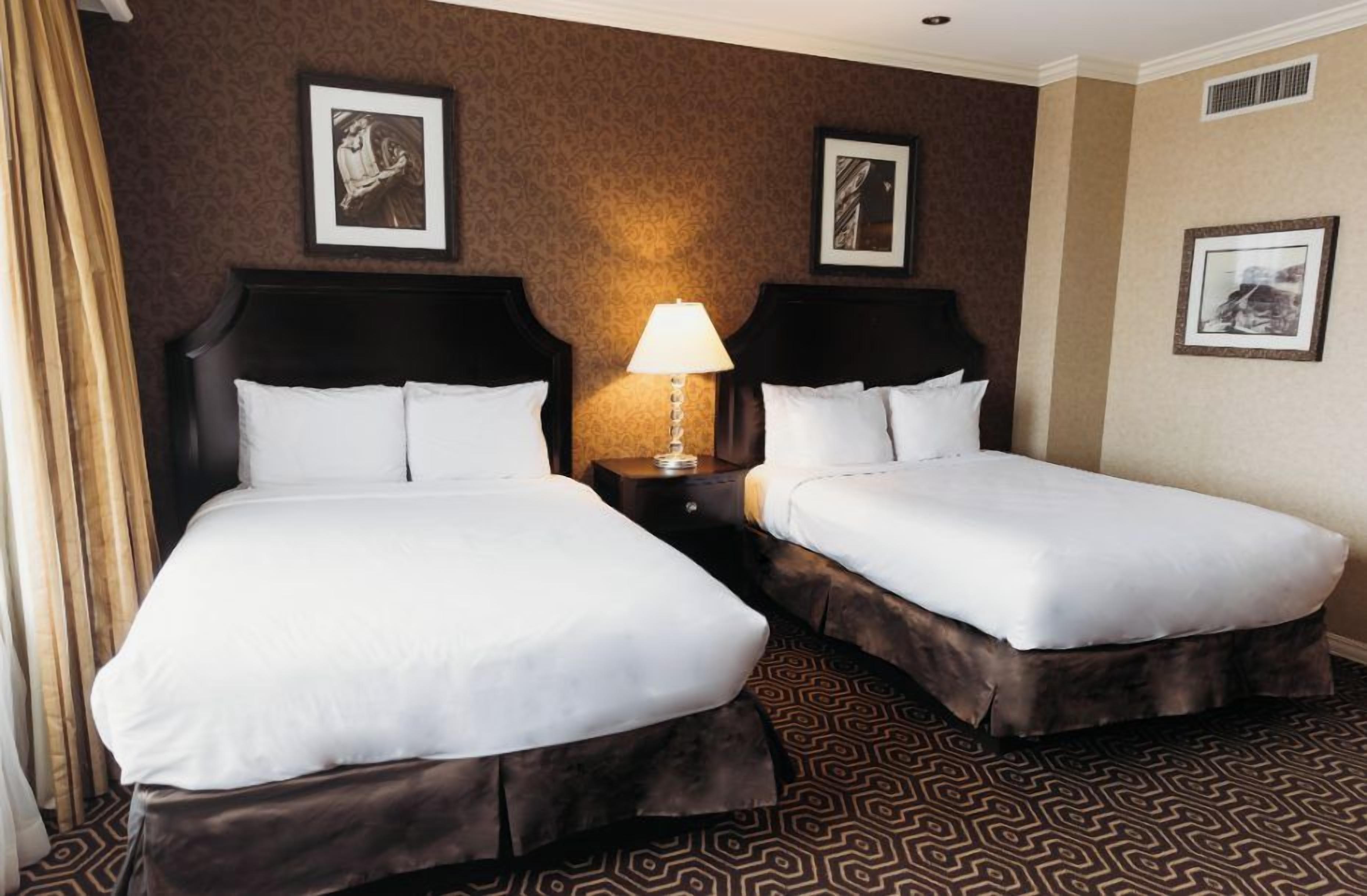 Habitación de hotel con dos camas, una mesita de noche con lámpara y alfombra estampada.