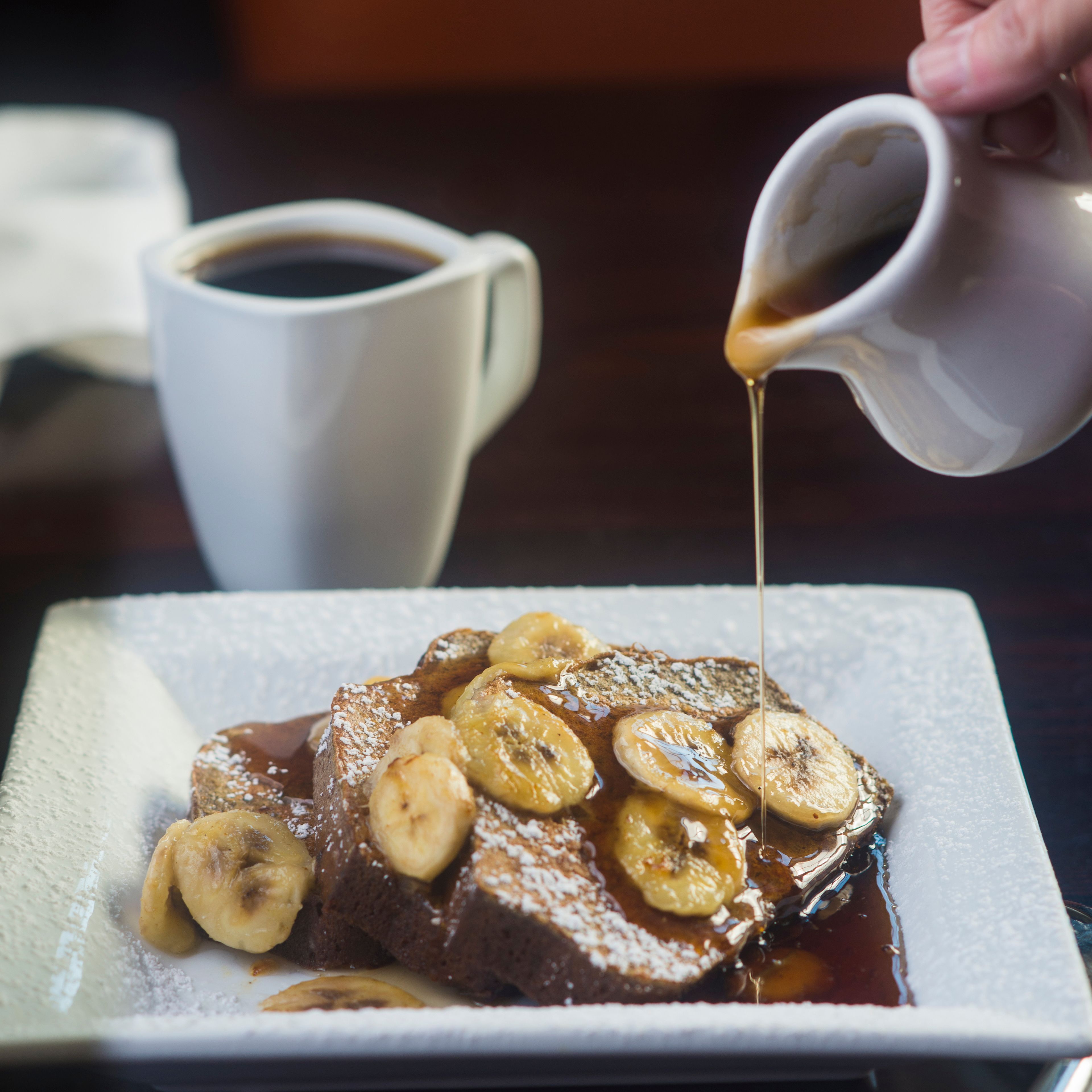 A plate of bread pudding topped with bananas and syrup, with a cup of coffee in the background.