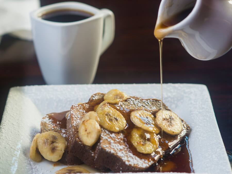 A plate of bread pudding topped with bananas and syrup, with a cup of coffee in the background.