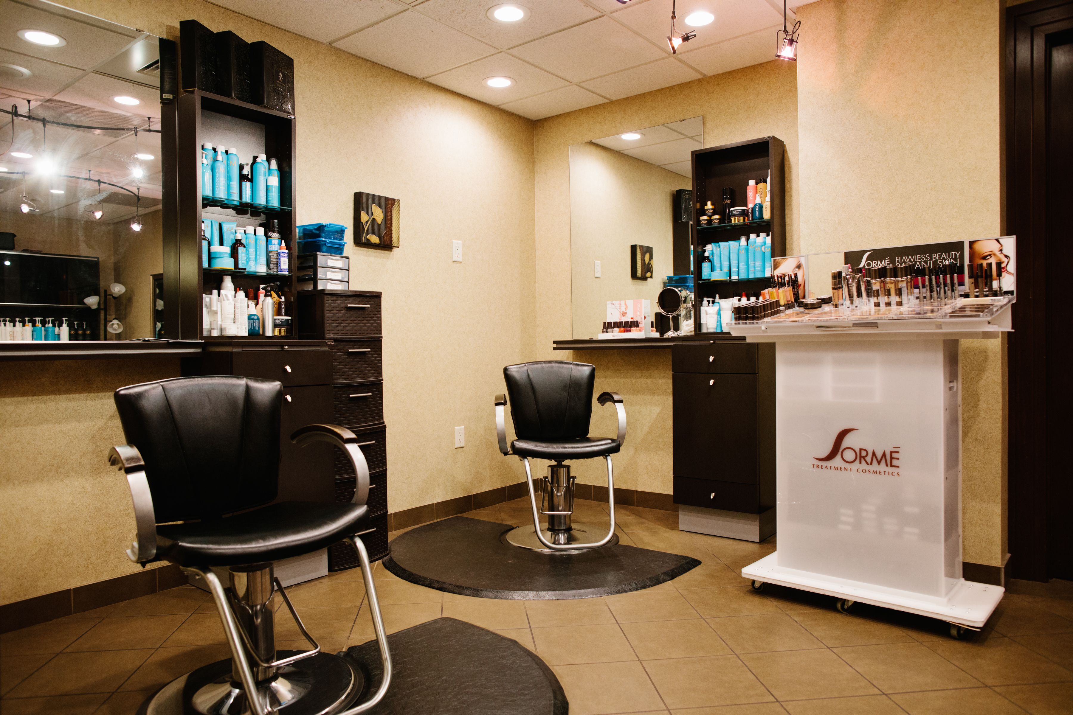 Een kapsalon met twee zwarte stoelen, een balie met diverse producten en een presentatie voor Sorme Treatment & Cosmetics.