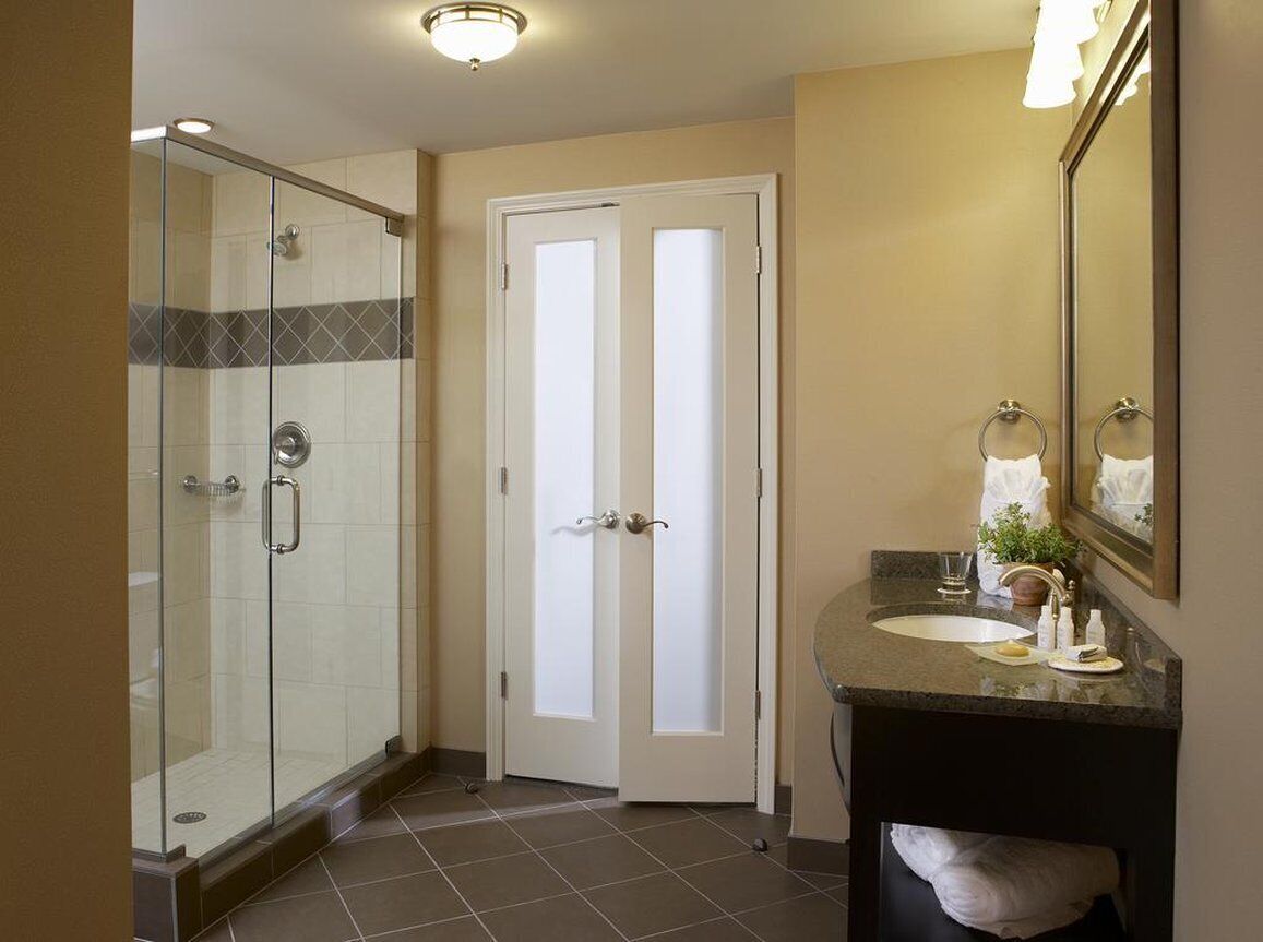 A bathroom with a glass shower enclosure, a white door, a sink with a mirror, and a towel rack.