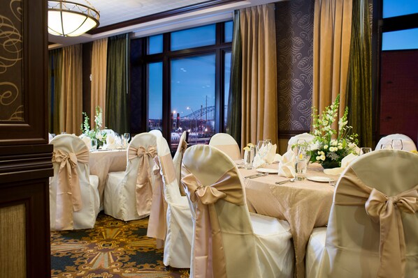 A banquet hall with round tables set for a formal event, large windows with a view of a bridge, and elegant decor.