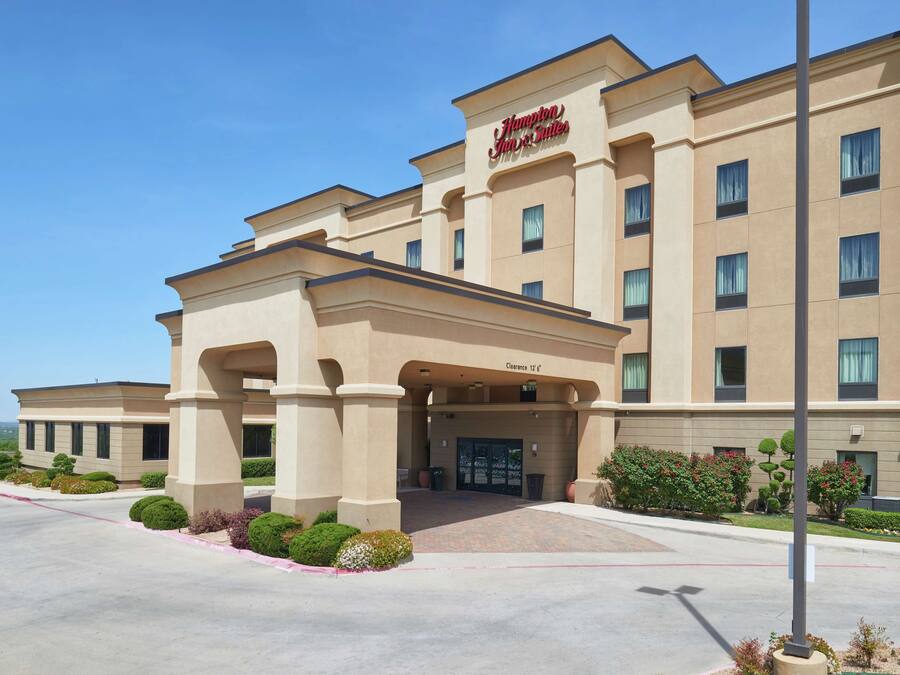 A Hampton Inn & Suites hotel with a beige facade, a central entrance with columns, and a clear sky.