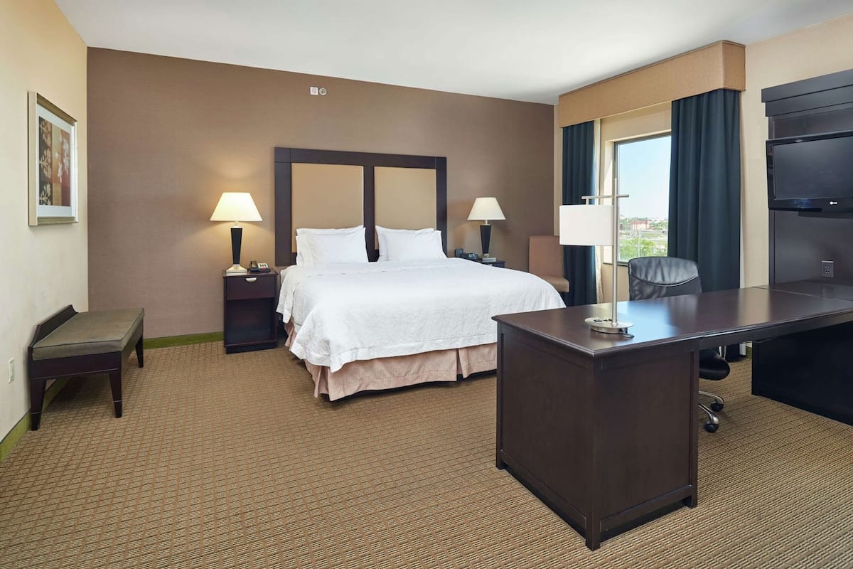 A hotel room with a bed, bedside tables, a desk, a chair, and a large window.