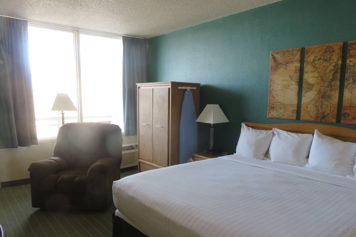 A hotel room with a large bed, a chair, a wardrobe, a lamp, and a window with curtains.