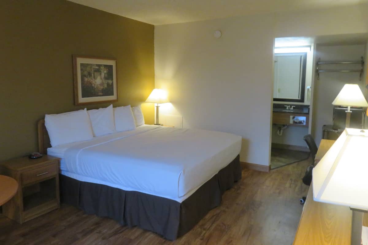 A hotel room with a bed, bedside lamps, a nightstand, a wooden floor, a painting on the wall, and a bathroom visible through an open door.